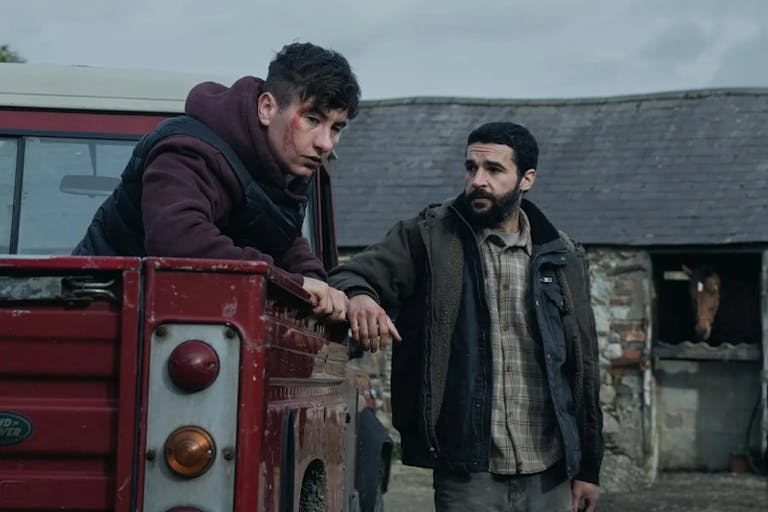 A young white man sitting in the back of a red truck with a cut on his head, a young Black man stands beside him looking contemplative