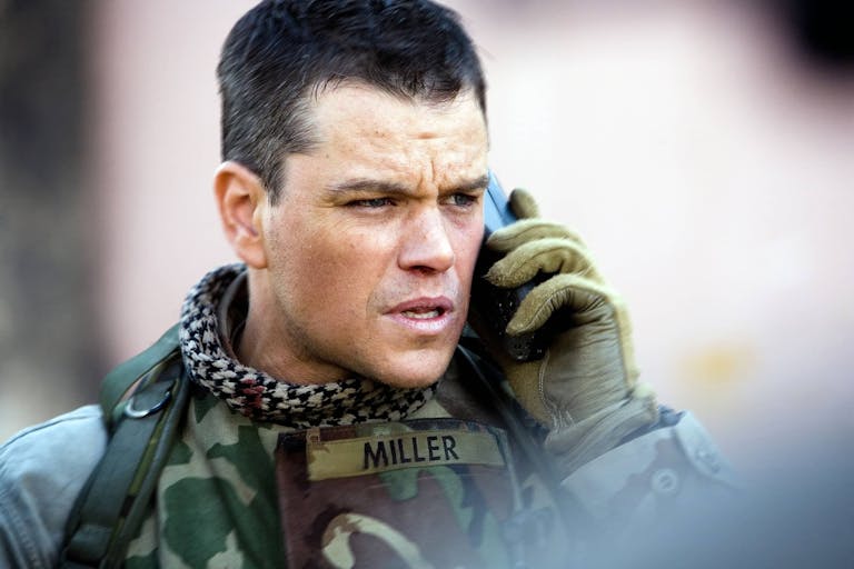 A white man in US army uniform and black and white keffiyeh holding a phone up to his ear 