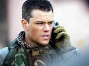A white man in US army uniform and black and white keffiyeh holding a phone up to his ear