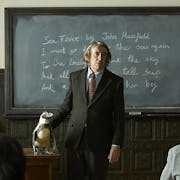A teacher, played by Steve Coogan, dressed in a dark suit with shirt and tie, stands in front of a classroom blackboard. Besides him is a wooden desk with a Magellanic penguin standing on it.