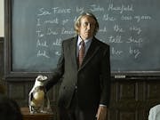 A teacher, played by Steve Coogan, dressed in a dark suit with shirt and tie, stands in front of a classroom blackboard. Besides him is a wooden desk with a Magellanic penguin standing on it.