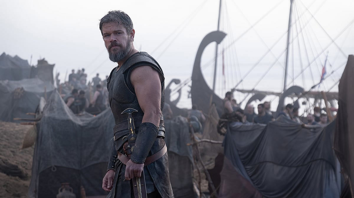 A man wearing brown ancient war armour, looking away with a concerned look on his face. Behind him, a large ship in the background