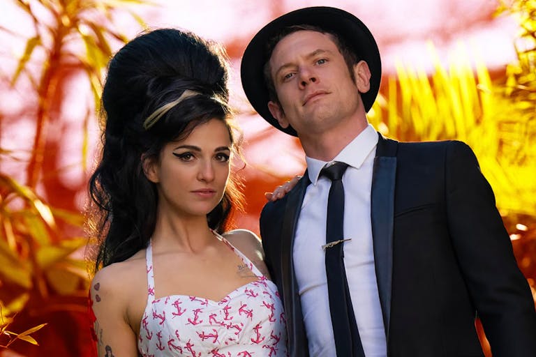 A young white woman with large beehive style hair and thickly flicked eyleliner and young white man in a suit and hat