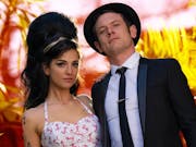 A young white woman with large beehive style hair and thickly flicked eyleliner and young white man in a suit and hat