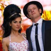 A young white woman with large beehive style hair and thickly flicked eyleliner and young white man in a suit and hat