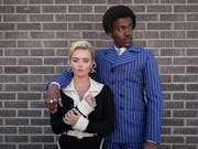 A young white woman and a young Black man in 60s fashionable clothing and hairstyle, his arm around her