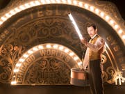 A young white man wearing a smart 1880's suit, holding a tube of light, with an arch of light bulbs over a sign reading 'TESLA'