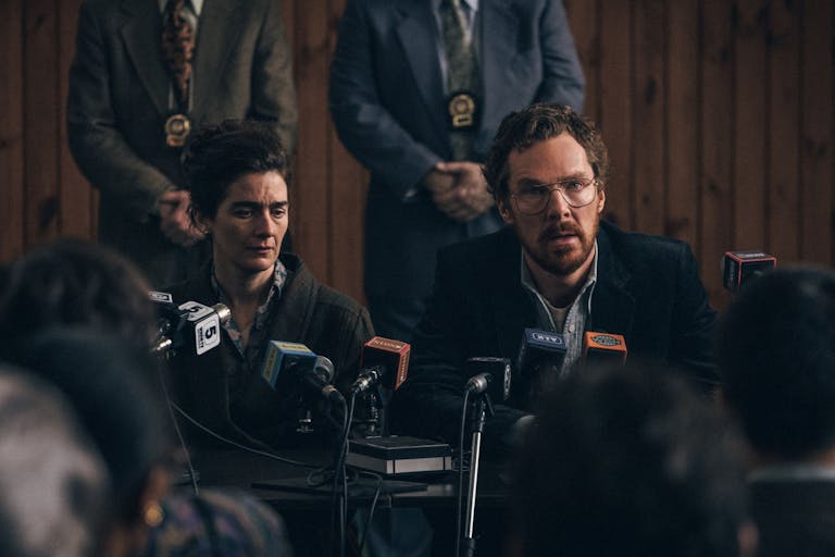A white woman and white man looking tired speaking at a press conference with police detectives standing behind them