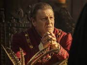 A confronted looking middle aged white man in regal red Tudor clothing and gold necklace
