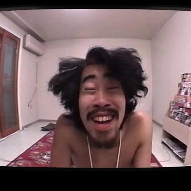 A Japanese man with wild dark hair in a room, staring manically at the camera, framed by a TV screen