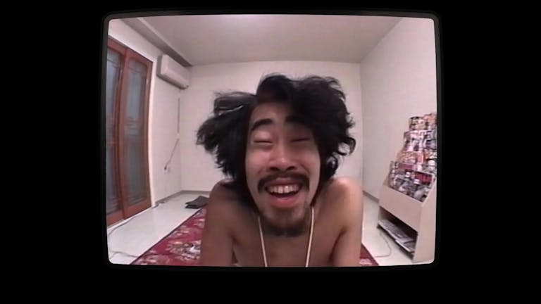 A Japanese man with wild dark hair in a room, staring manically at the camera, framed by a TV screen