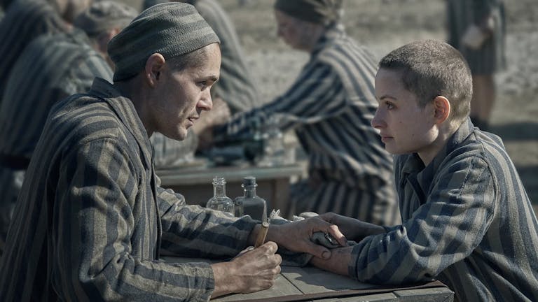 A white man and white woman with shaved heads in striped concentration camp clothing looking across as each other
