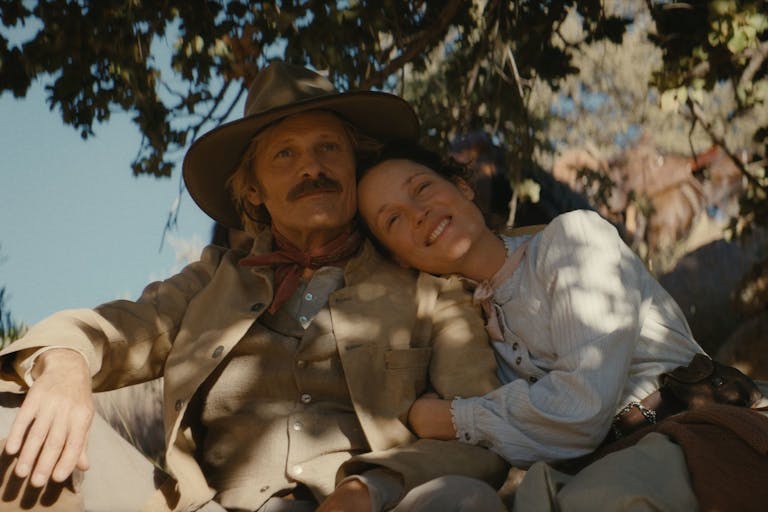 A white man and white woman in traditional 19th Century western clothing sitting under the shade of a tree, hugging and looking content