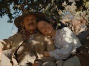 A white man and white woman in traditional 19th Century western clothing sitting under the shade of a tree, hugging and looking content