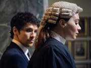 Side profile of a young white woman barrister in robes and wig, as a young white man looks across at her