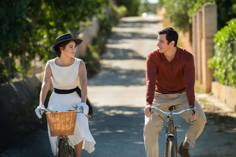 A young white woman and young white man in 60s clothing, cycling in the sun smiling at each other
