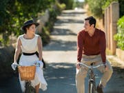 A young white woman and young white man in 60s clothing, cycling in the sun smiling at each other