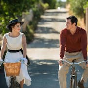 A young white woman and young white man in 60s clothing, cycling in the sun smiling at each other