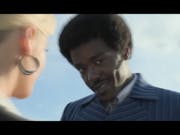A young Black man in a pin stripe suit and short afro smiles coyly at the camera