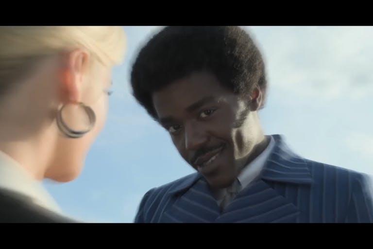 A young Black man in a pin stripe suit and short afro smiles coyly at the camera