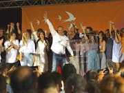 A large group of people on stage cheering and holding their arms up in triumph and peace signs