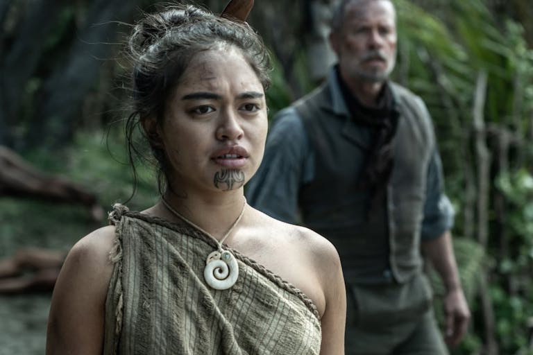 A young Māori woman in indigenous clothing with a chin tattoo stands in forest with a older white man standing in the background