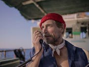 A middle aged white man wearing a red hat wearing rugged blue shirt and handkerchief around his neck, speaking into a radio
