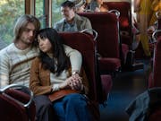 A young white man and young Japanese woman sit cuddling on a bus looking sorrowful
