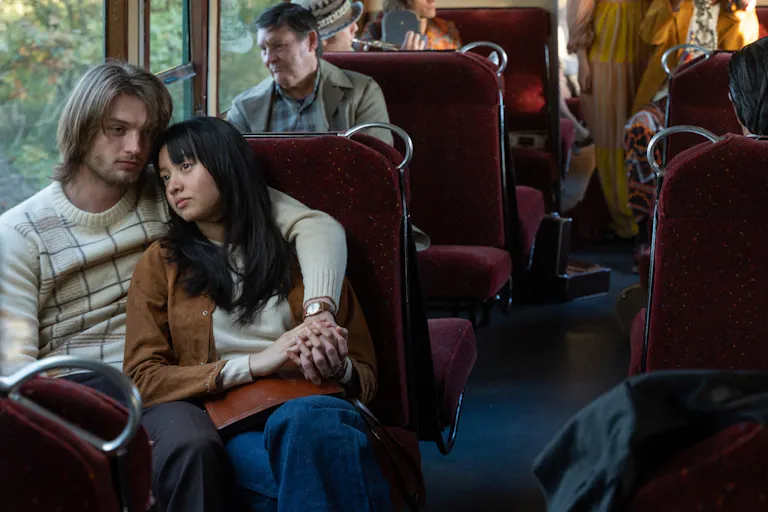 A young white man and young Japanese woman sit cuddling on a bus looking sorrowful