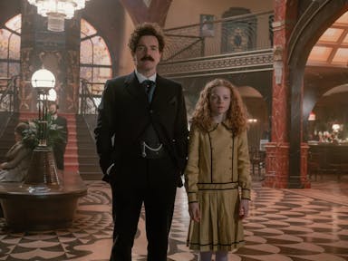 A white man with short, dark curly hair and young white girl with curly orange hair stand in an opulent, grand, art deco foyer