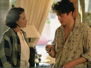 A white woman and man in 90s casual clothing stand facing each other discussing a watch in his hand
