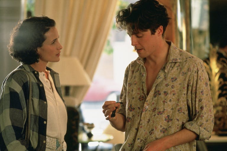 A white woman and man in 90s casual clothing stand facing each other discussing a watch in his hand
