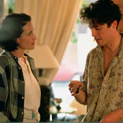 A white woman and man in 90s casual clothing stand facing each other discussing a watch in his hand