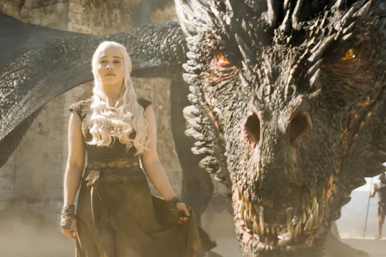 A young white man with bright white hair stands defiant as a huge, scaled dragon with amber eyes crouches behind