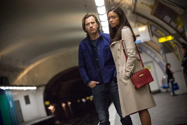 A white man and a Black woman stand on the edge of a train platform, looking serious