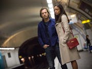 A white man and a Black woman stand on the edge of a train platform, looking serious