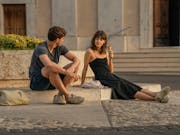 A young white man and young white woman sit in a stone European square eating ice cream, looking at each other