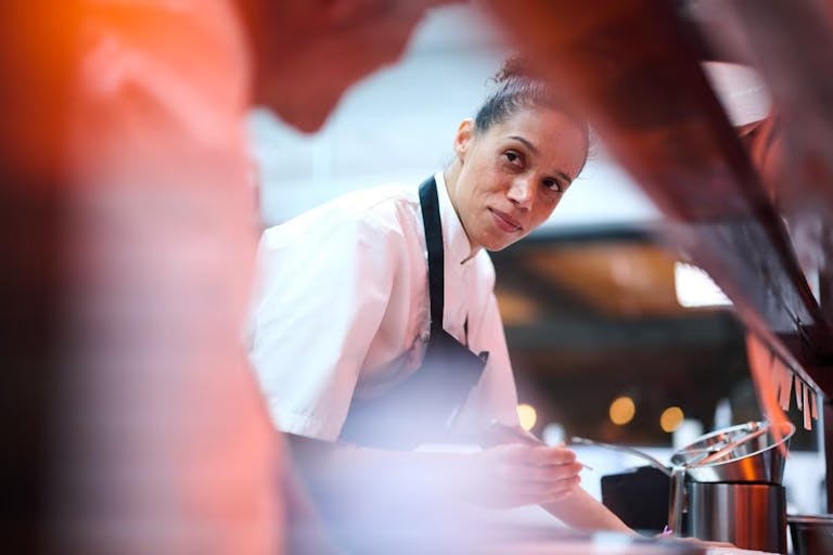 A mixed heritage Black woman with hair tied back in a bun wearing chef whites and an apron in a restaurant kitchen