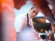 A mixed heritage Black woman with hair tied back in a bun wearing chef whites and an apron in a restaurant kitchen