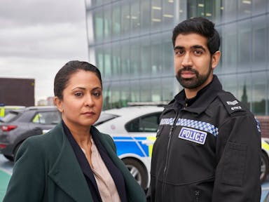 A Brown woman detective and a young brown policeman in uniform standing in front of a police car