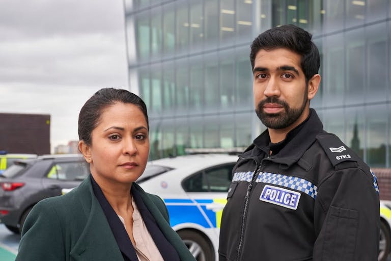 A Brown woman detective and a young brown policeman in uniform standing in front of a police car