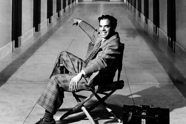 Black and white image of a white man sat in a folding directors chair pointing down a hall
