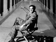 Black and white image of a white man sat in a folding directors chair pointing down a hall