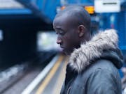 A young Black teen boy in a parka standing on the edge of a tube platform looking scared and sad