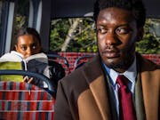 A Black man in a suit and tie sits on a bus as a young Black woman sits a seat behind him looking at him over her book