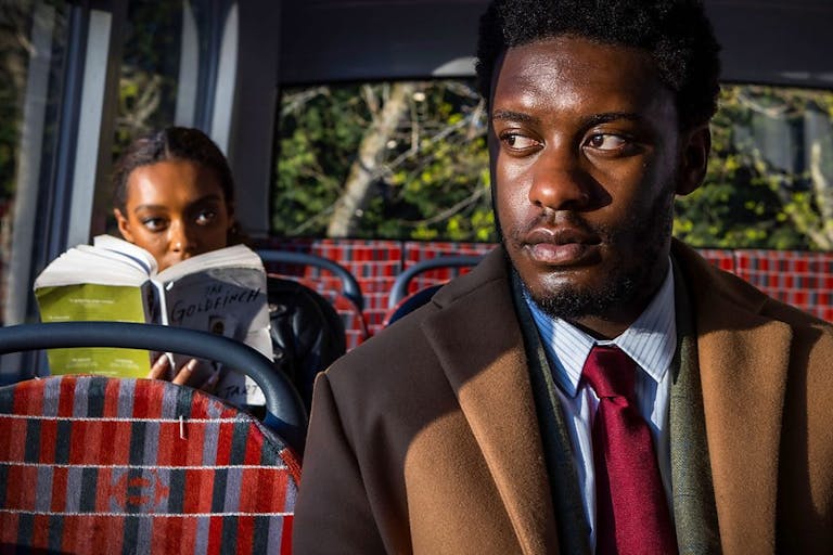 A Black man in a suit and tie sits on a bus as a young Black woman sits a seat behind him looking at him over her book