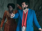 A Black woman in orange velvet corset dress and a white man with large mutton chops and wearing a blue jacket and red waistcoat, pointing at something