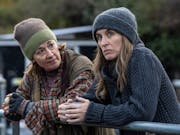 Two white women in jumpers and woolly hats holding a mug, leaning over a metal farm fence, looking serious