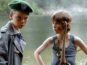 Two young white boys, one in an oversized army cadet uniform and beret, and the other in a grey vest, black stripes across his cheeks and fabric tied around his head