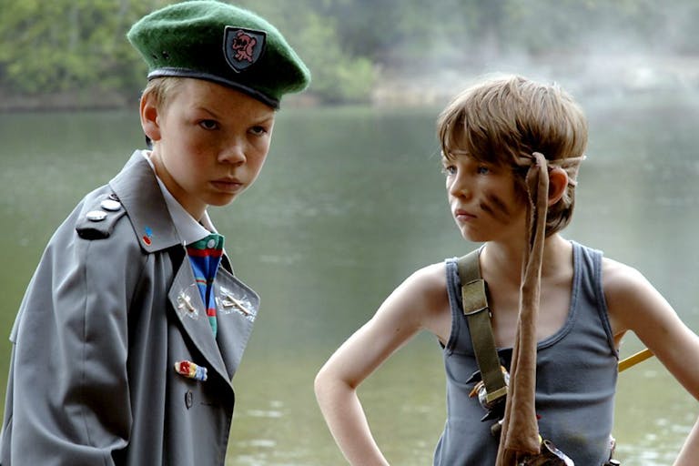 Two young white boys, one in an oversized army cadet uniform and beret, and the other in a grey vest, black stripes across his cheeks and fabric tied around his head